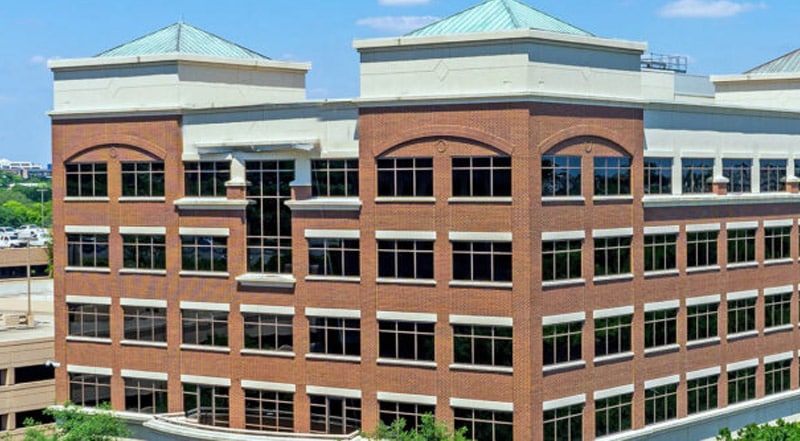 A multi-story office building with red brick walls, large windows, and greenish metal roofing, seen on a sunny day with a blue sky and some trees in front.