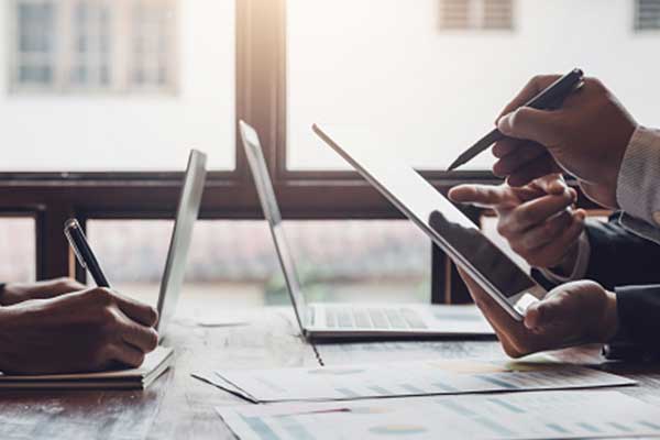 People working together at a table with laptops and documents. One person writes on paper, another holds a tablet, and a third gestures with a pen, suggesting a business meeting or collaborative discussion.
