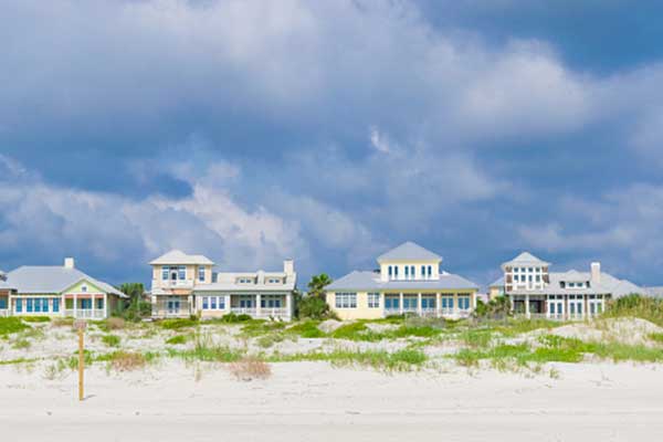 Several beach houses with light-colored exteriors sit behind sand dunes and grass under a cloudy sky.