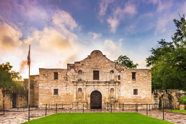 The Alamo mission in San Antonio, Texas, is shown at sunrise with a cloudy sky, stone facade, arched doorway, and a flagpole on the left, surrounded by trees and green grass in the foreground.