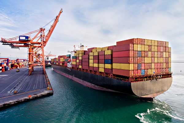 A large cargo ship loaded with multicolored shipping containers is docked at a port, with cranes and more containers visible on the dock under a cloudy sky.