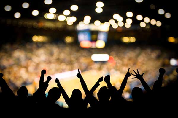 Silhouetted crowd cheering with raised arms at an indoor sports event, with a brightly lit court and out-of-focus audience in the background.