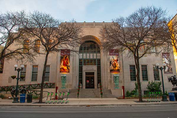 A historic stone building with tall arched entrance, flanked by leafless trees and colorful banners. Electric scooters are lined up on the sidewalk in front of the building.