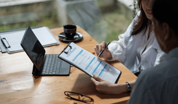 Two people sit at a wooden table, reviewing and filling out a form on a clipboard. A tablet with a keyboard, a pen, and a cup of coffee are also on the table. The setting appears to be casual and professional.
