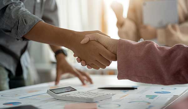 Two people shake hands over a desk with documents and a calculator, suggesting a business agreement or partnership. Other blurred figures stand in the background.