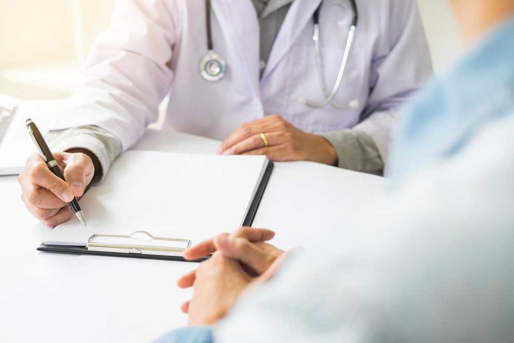A doctor in a white coat writes on a clipboard while speaking with a patient, whose hands are folded on the table.