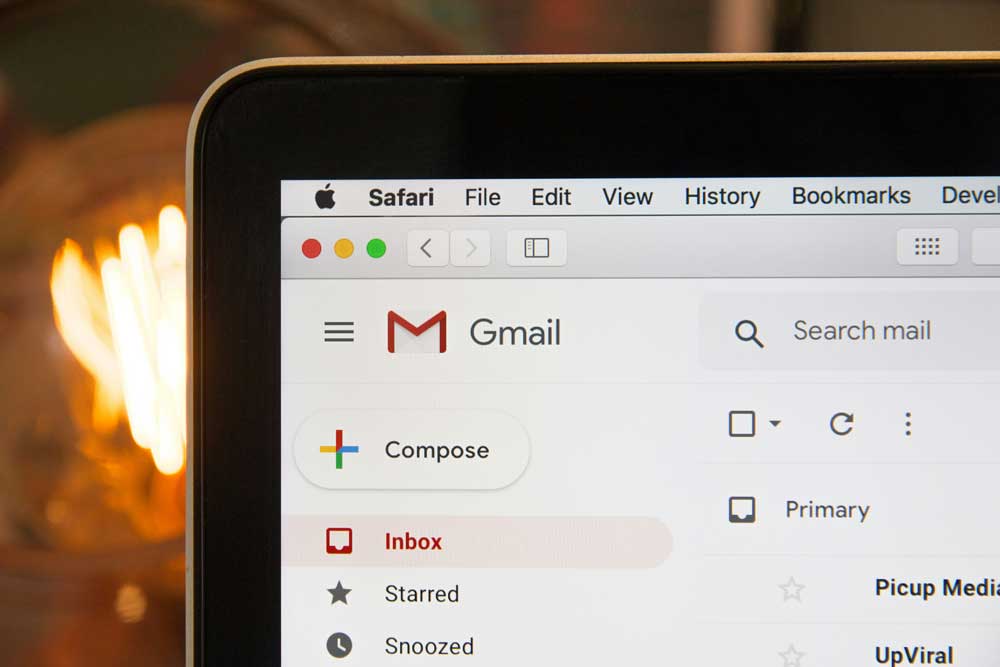 Close-up of a laptop screen showing the Gmail interface, including the Compose button, inbox, and mail categories. The Safari browser tab and menu bar are visible at the top.