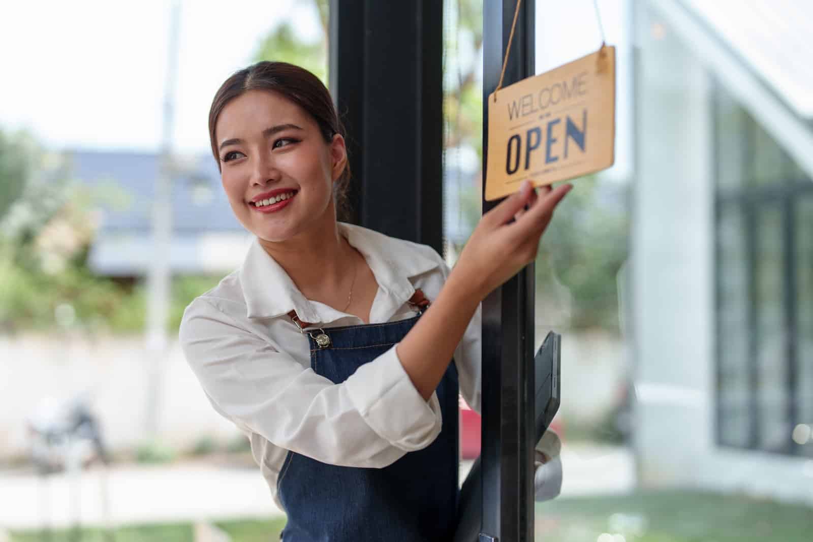 A smiling cafe worker in an apron turns a sign on a glass door that reads Welcome Open.